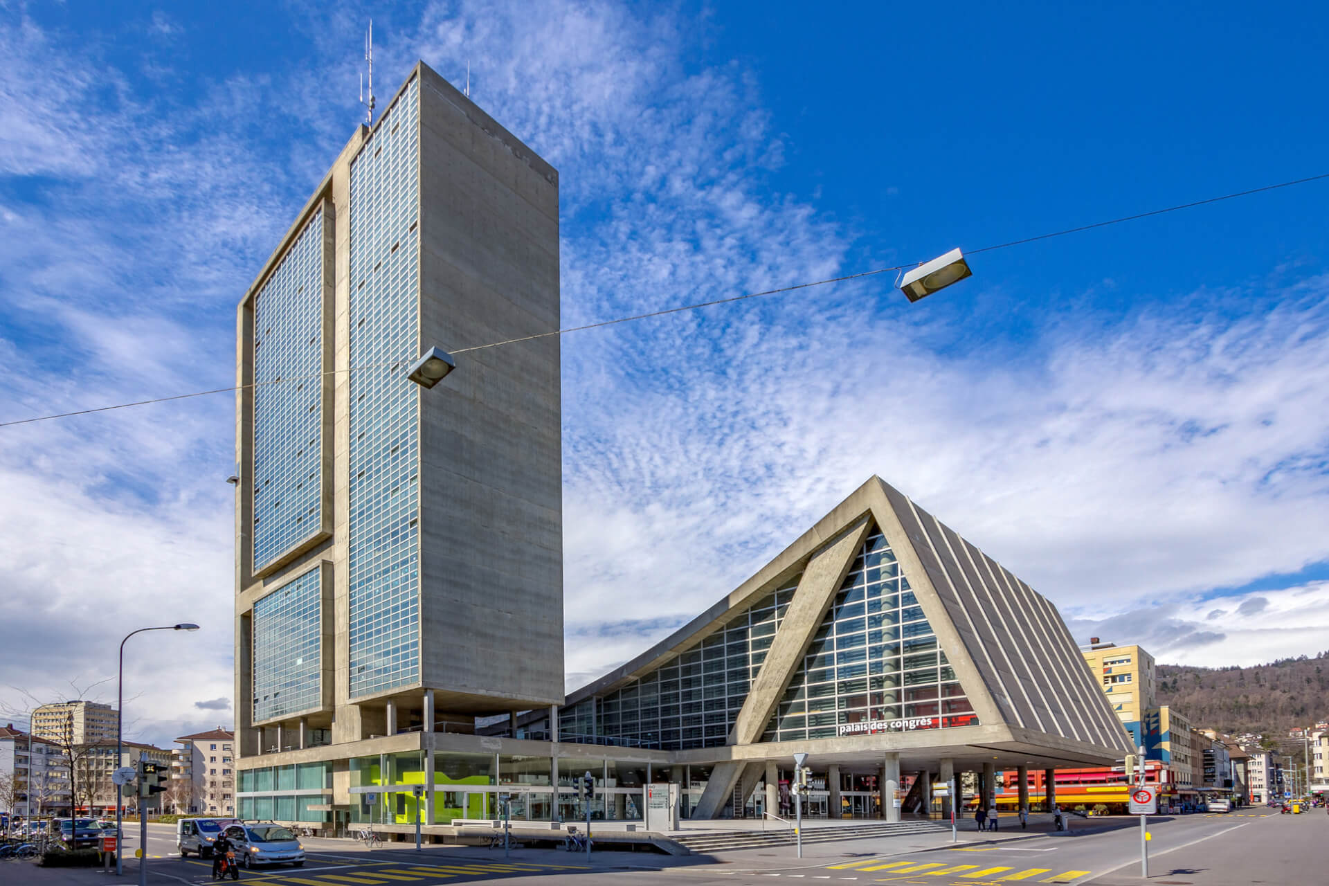 Bâtiment moderne avec une haute tour rectangulaire et une structure de verre triangulaire adjacente, situé dans une rue de la ville sous un ciel bleu avec des nuages épars.