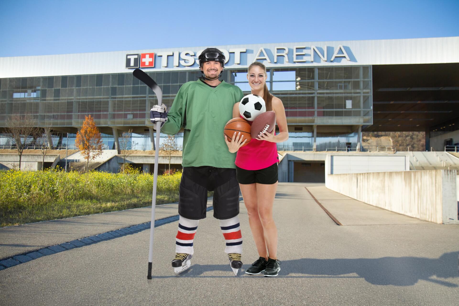 Ein Eishockeyspieler in voller Montur und eine Frau in Sportkleidung, die einen Fußball und einen Basketball hält, stehen vor der Tissot Arena.