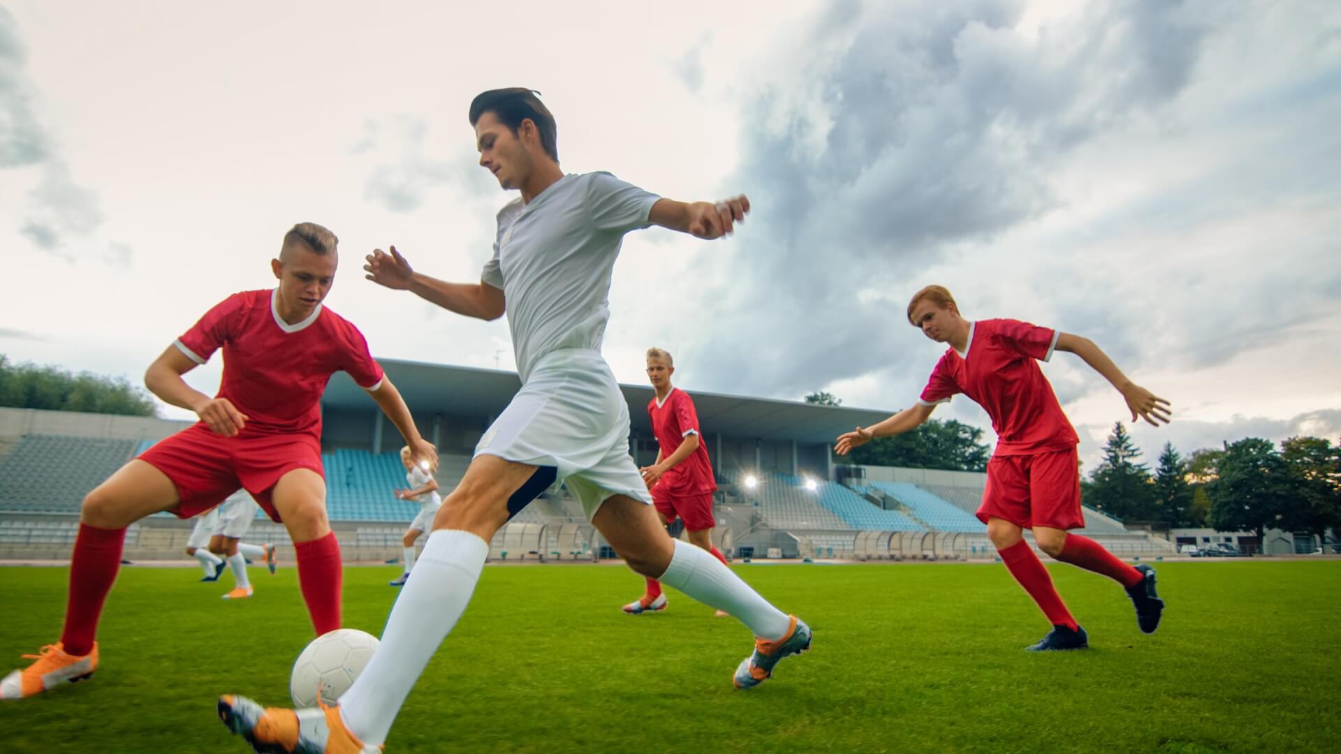 Un joueur de football en tenue blanche dribble le ballon tandis que trois adversaires en tenue rouge tentent de le défendre sur un terrain en plein air.