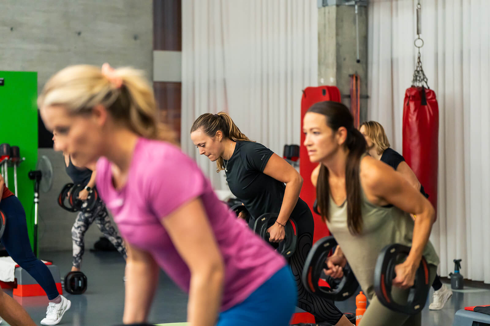 Un groupe de femmes en tenue de sport soulève des poids lors d'un cours de fitness dans une salle de sport, avec des équipements visibles à l'arrière-plan.