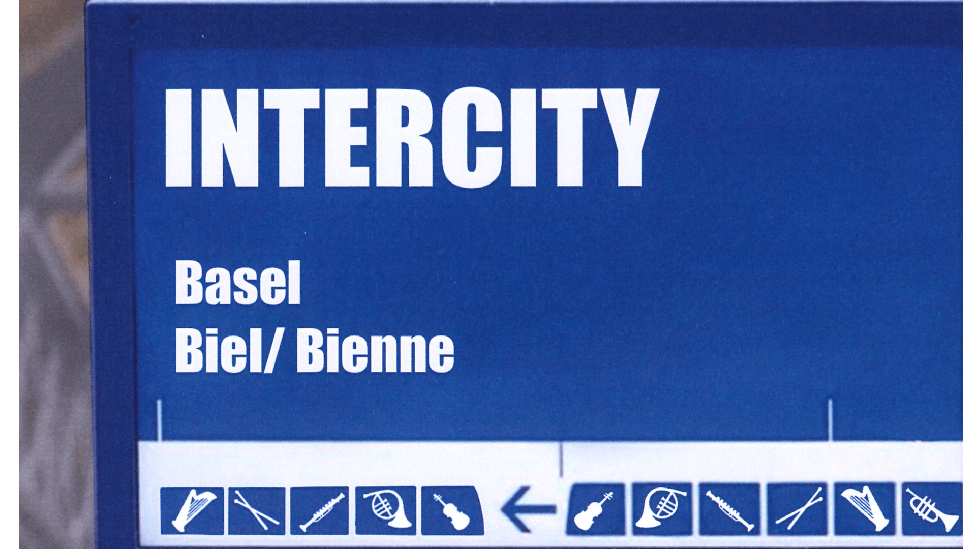 Blaues Schild mit der Aufschrift INTERCITY, mit den Zielorten Basel und Biel/Bienne und Symbolen von Musikinstrumenten am unteren Rand.