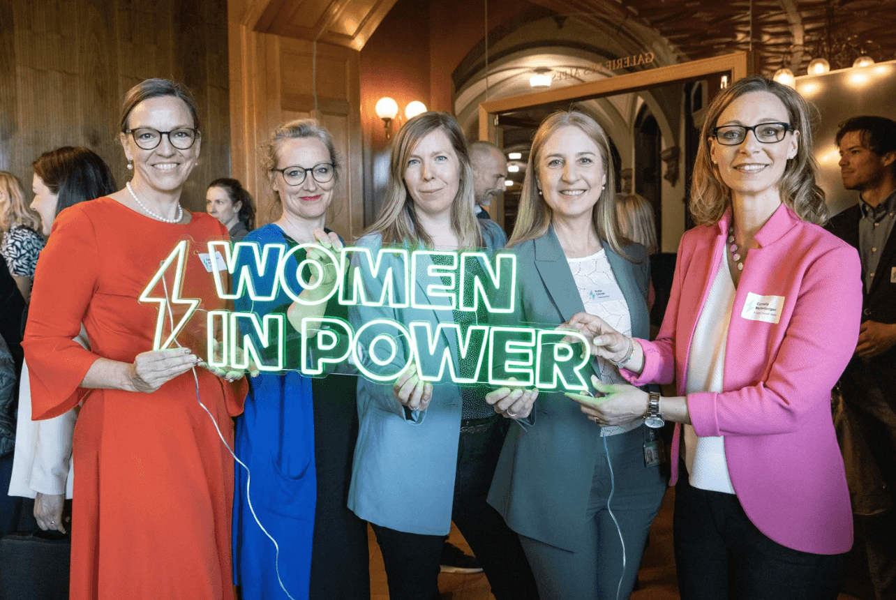 Fünf Frauen stehen in einem Innenraum zusammen, lächeln und halten ein Leuchtreklame-Schild mit der Aufschrift 