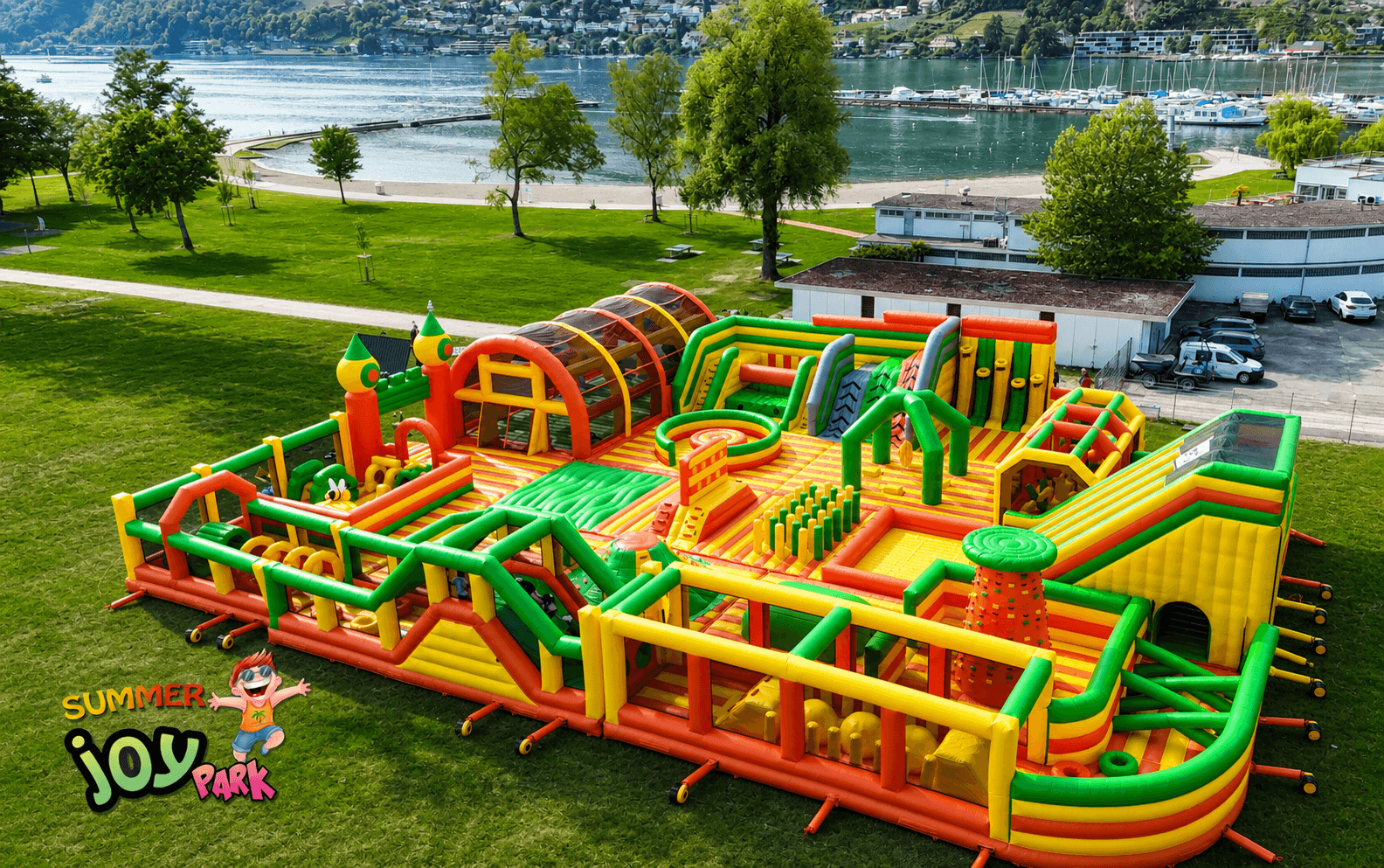 Großer aufblasbarer Hindernisparcours in leuchtenden grünen, gelben und orangenen Farben auf einer Wiese in der Nähe eines Sees und eines Jachthafens, mit Bäumen und Gebäuden im Hintergrund. Summer Joy Park-Logo in der Ecke.