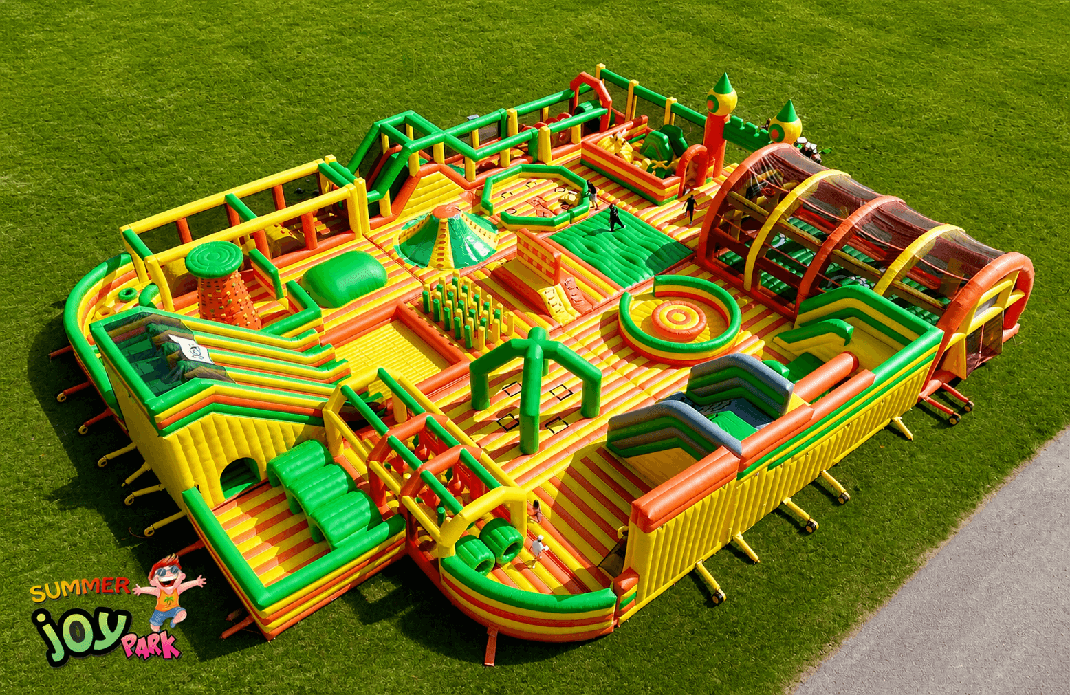 Ein großer, farbenfroher aufblasbarer Hindernisparcours und Spielplatz ist auf einer Wiese aufgebaut. Die Struktur verfügt über Rutschen, Kletterbereiche und Tunnels. SUMMER joy PARK ist in einer Ecke aufgedruckt.