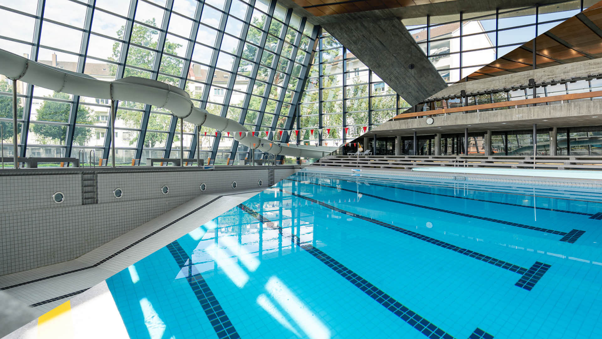 Ein Hallenbad mit klarem, blauem Wasser, freien Bahnen, einer Wasserrutsche und großen, schrägen Fenstern, durch die Tageslicht hereinströmt.