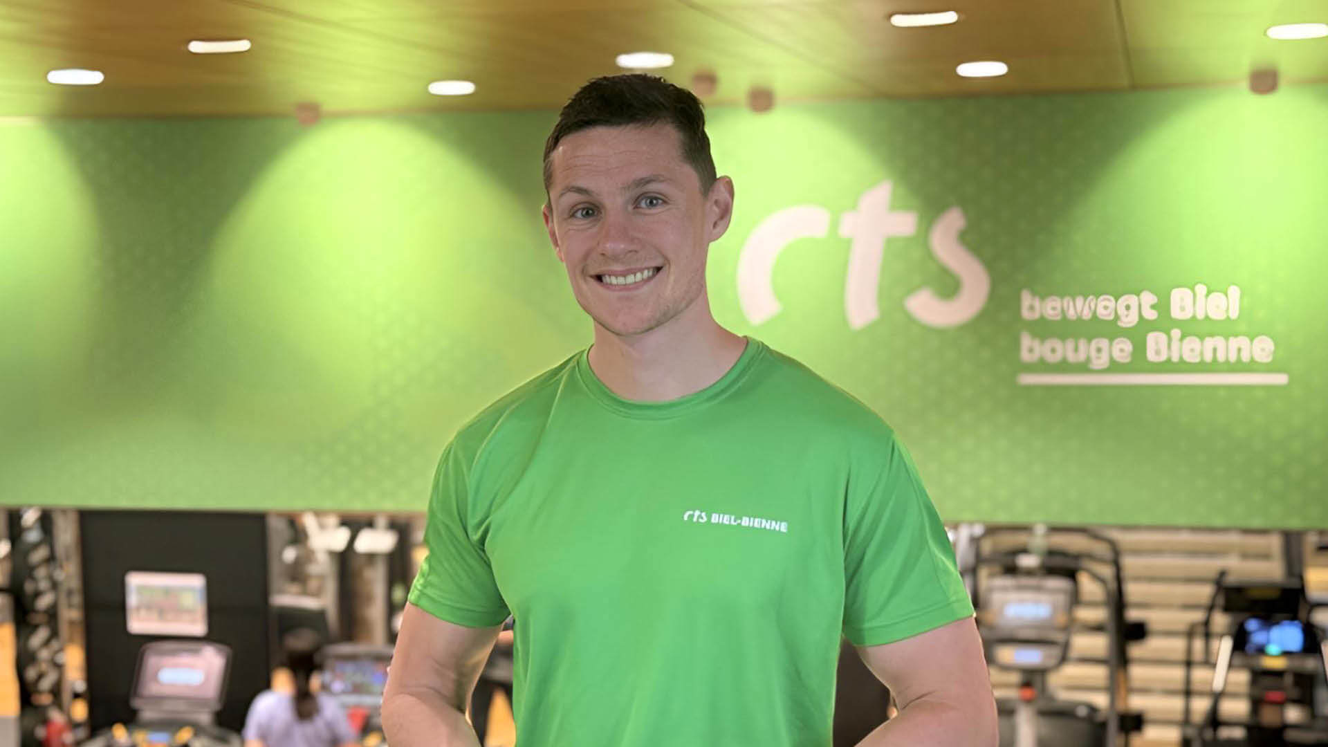 Une personne vêtue d'une chemise verte du CTS Biel-Bienne sourit dans une salle de gymnastique, avec des appareils d'exercice visibles en arrière-plan.
