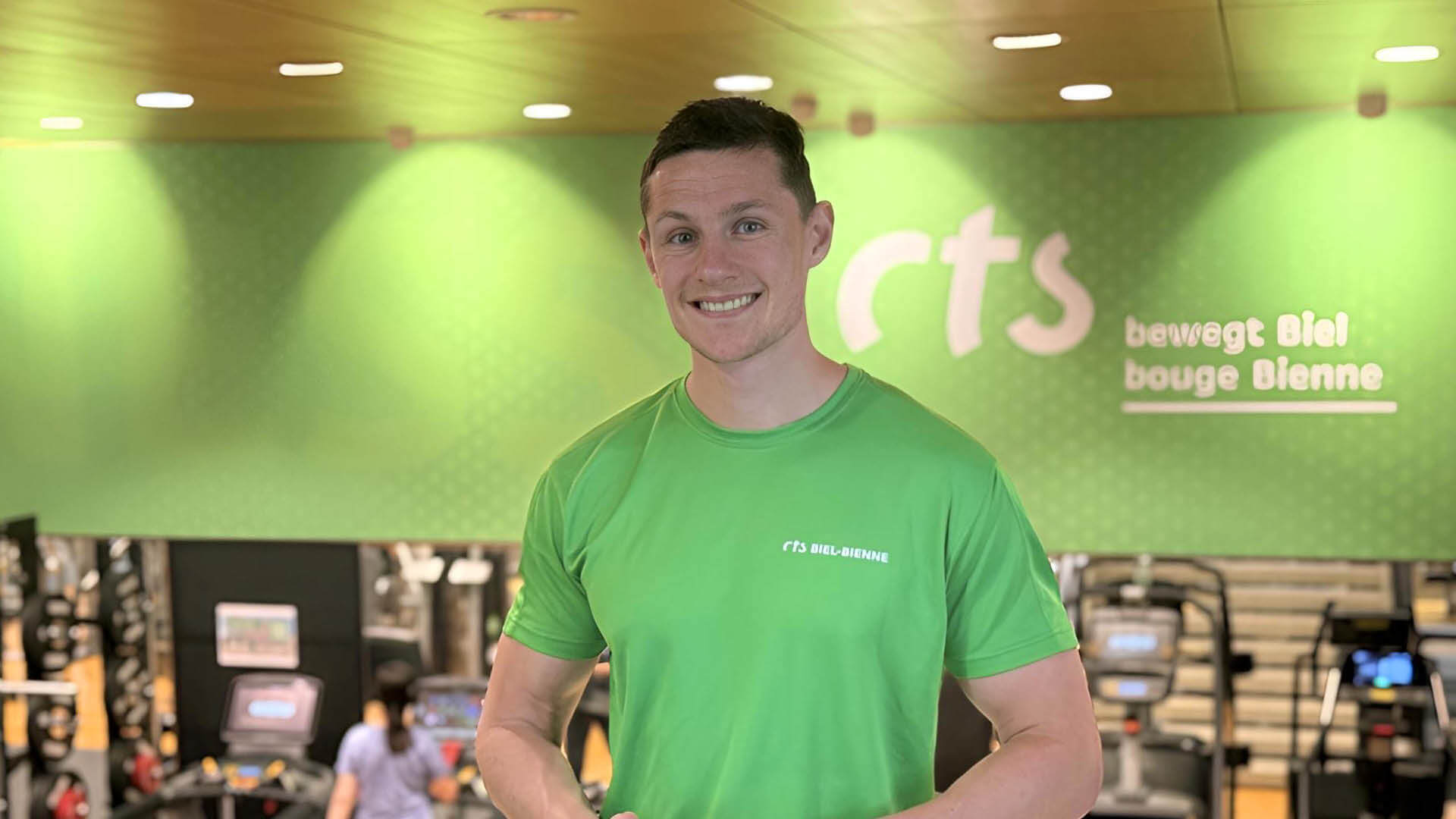 Ein Mann in einem grünen T-Shirt steht lächelnd in einem Fitnessstudio mit Fitnessgeräten und einer grünen Wand mit 