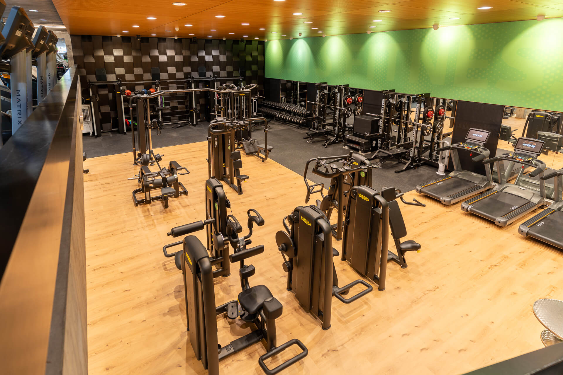 Geräumiger Fitnessraum mit verschiedenen Trainingsgeräten, Hanteln und Laufbändern auf einem Holzboden, unter einer gemusterten Decke und einer Wand mit grünen Akzenten.