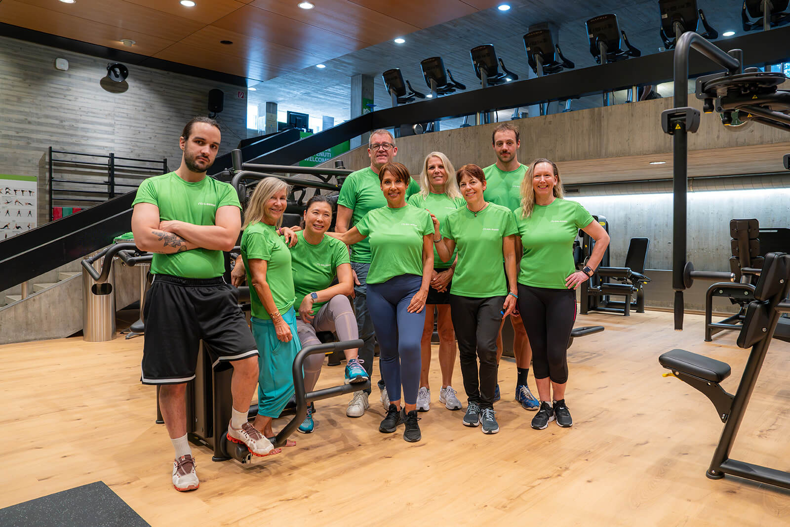 Neuf personnes vêtues de t-shirts verts identiques posent ensemble dans une salle de sport moderne, debout au milieu d'appareils de fitness posés sur un parquet, avec des escaliers et des murs en béton en arrière-plan.