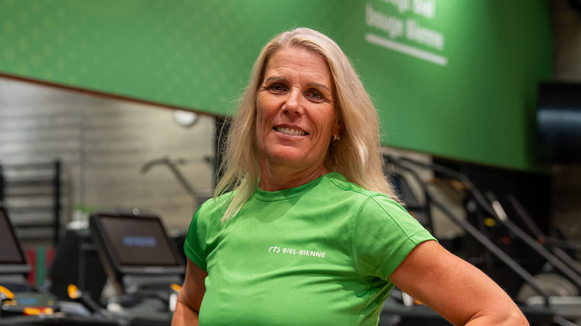 Eine Frau mit langen blonden Haaren, die ein grünes Shirt trägt, steht in einem Fitnessstudio und posiert lächelnd vor Laufbändern und einer grünen Wand.
