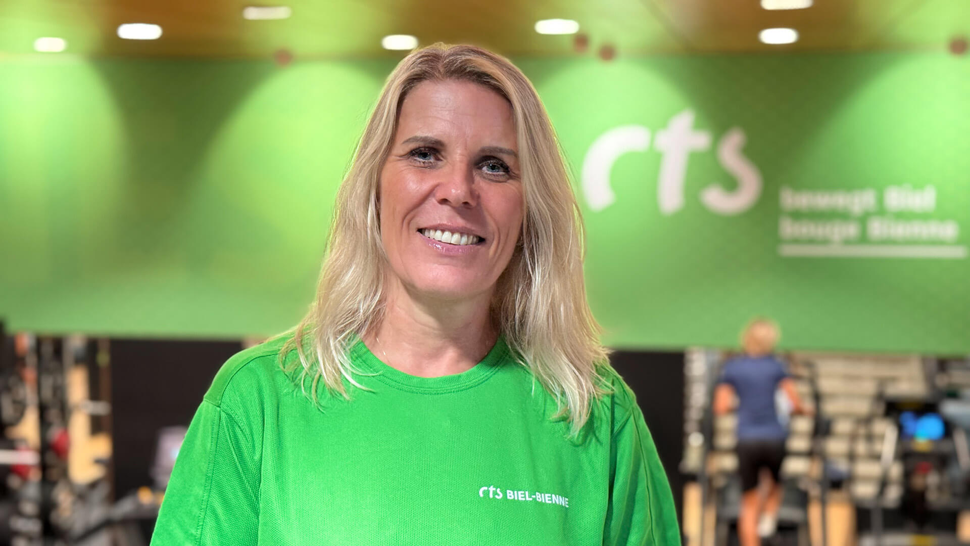 Eine Frau in einem grünen „rts BIEL-BIENNE“-Shirt lächelt in ein Kameraobjektiv, im Hintergrund sind ein Fitnessstudio mit Trainingsgeräten und eine grüne Wand zu sehen.