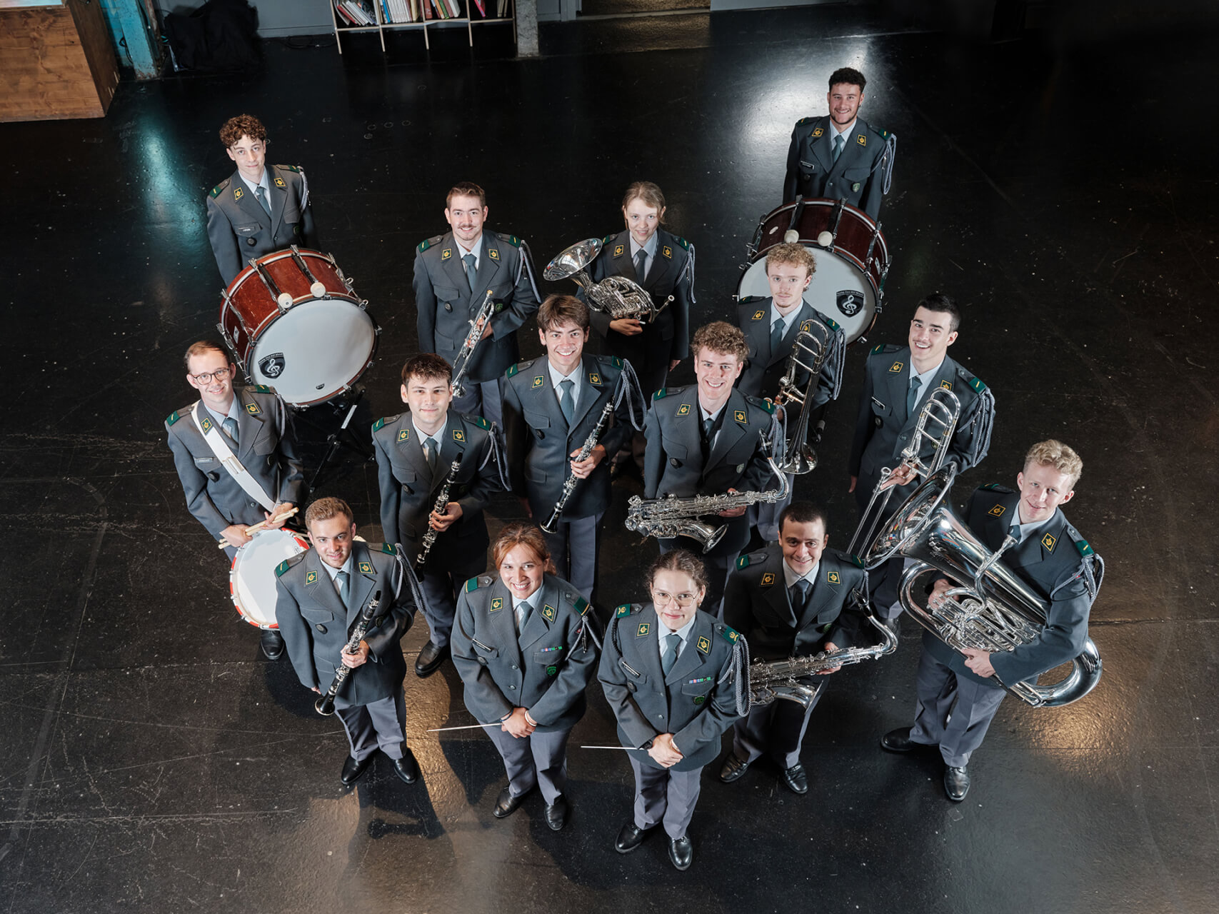 Eine Gruppe von vierzehn Personen in einheitlichen Uniformen, von denen jede ein Musikinstrument in der Hand hält, posiert gemeinsam auf einem dunklen Boden und blickt in die Kamera.