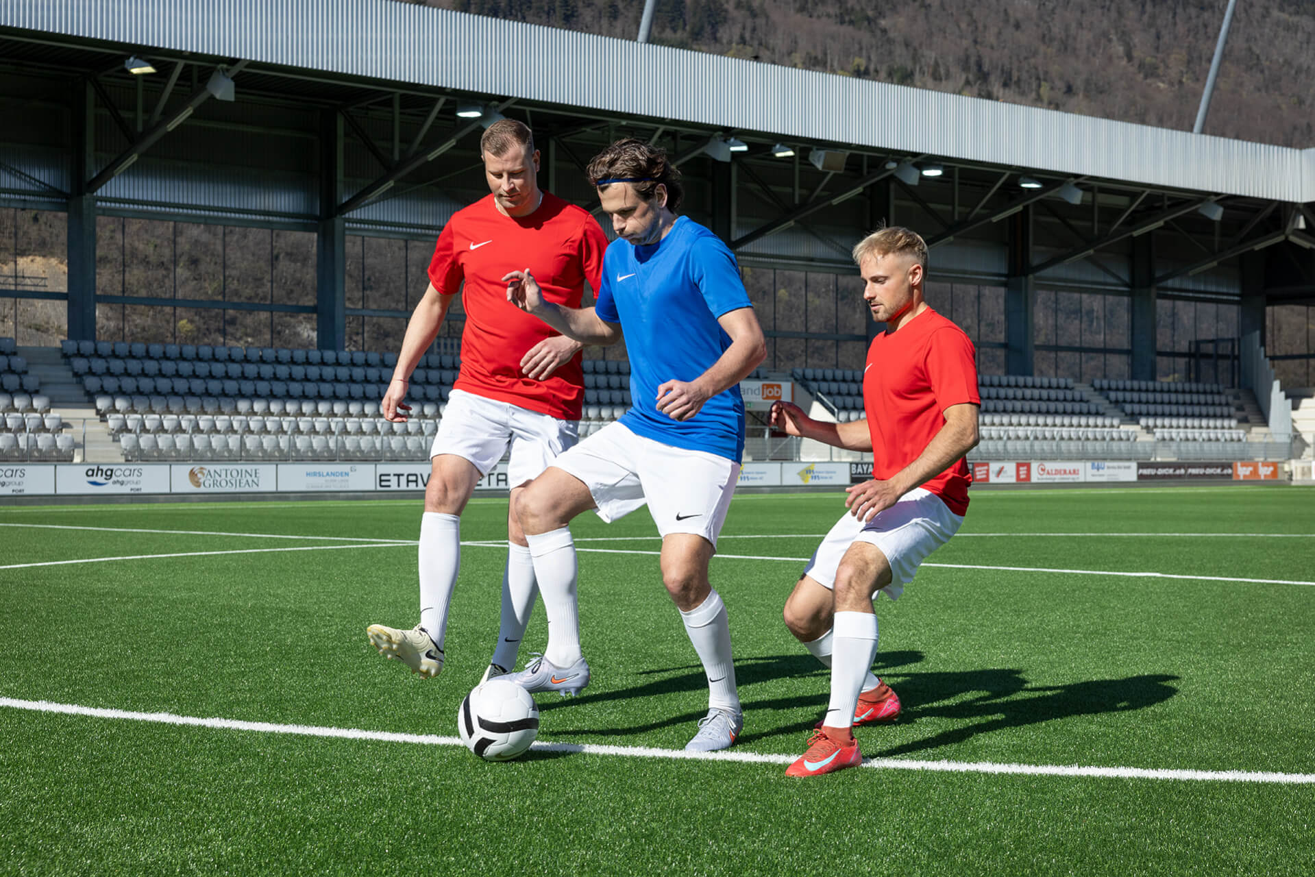 Trois hommes adultes jouent au football sur un terrain en plein air. Deux d'entre eux, vêtus de maillots rouges, défendent face à un troisième, en maillot bleu, qui a le ballon. On aperçoit des sièges vides dans les tribunes, à l'arrière-plan.