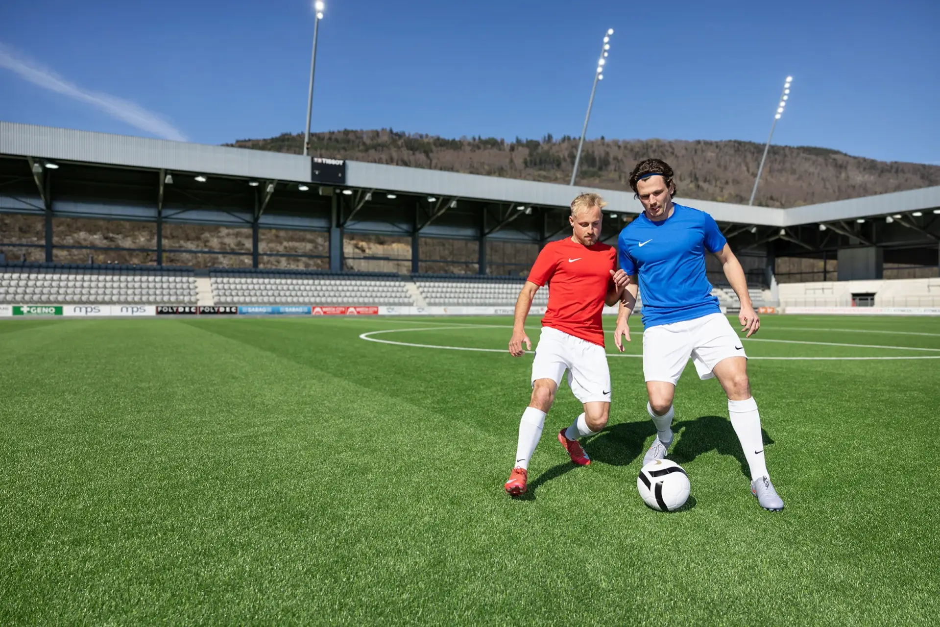 Deux footballeurs, vêtus de maillots rouges et bleus, se disputent le ballon sur un terrain vert, dans un stade en plein air, sous un ciel dégagé.