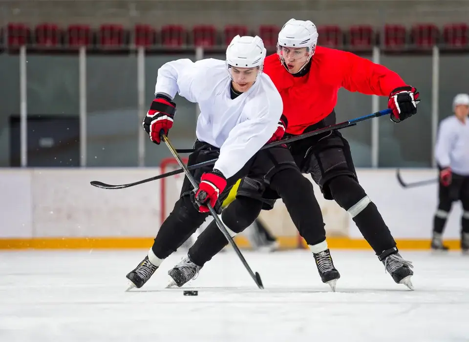 Deux joueurs de hockey sur glace en tenue complète se disputent le palet sur une patinoire couverte ; l'un est vêtu de blanc, l'autre de rouge, tandis que deux autres joueurs sont visibles à l'arrière-plan.