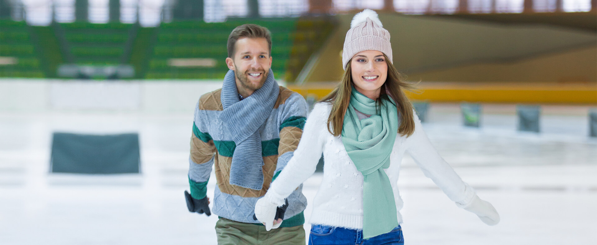 Un homme et une femme, vêtus de vêtements d'hiver, font du patin à glace dans une salle, main dans la main, tout en souriant.