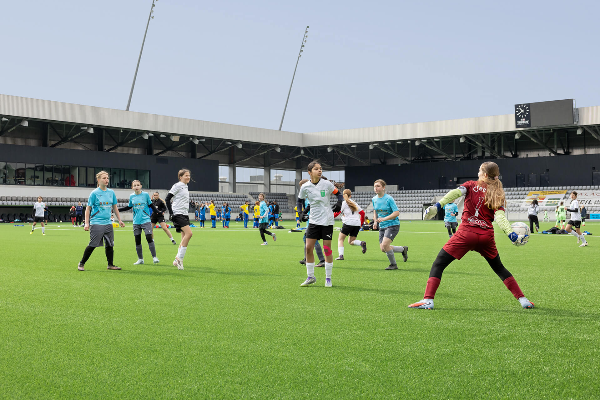 Un gardien de but en rouge s'apprête à relancer le ballon tandis que des joueurs vêtus de maillots blancs et bleu sarcelle s'affrontent sur un terrain de football dans un stade.