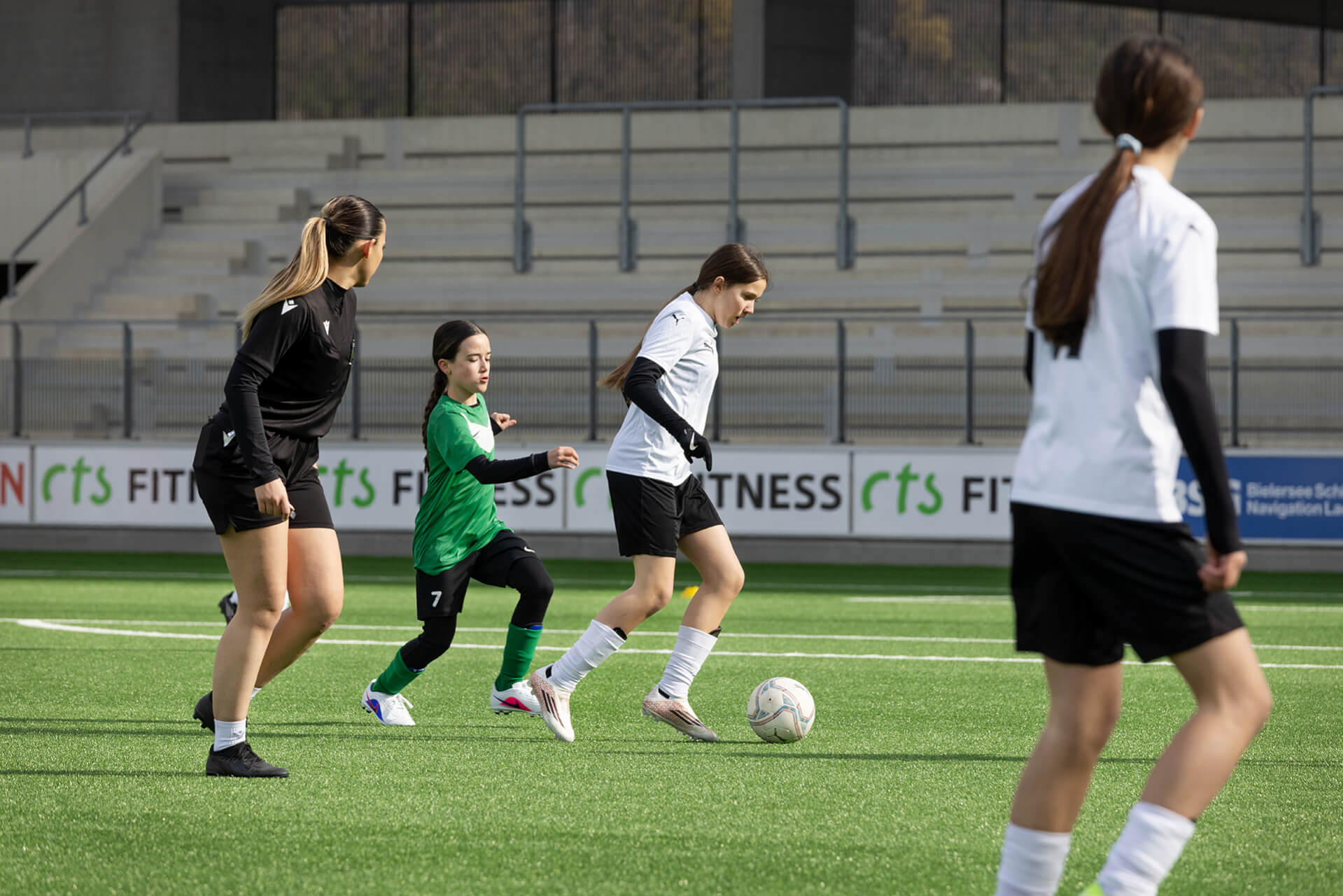 Quatre filles jouent au football sur un terrain vert ; l'une d'elles, vêtue d'un maillot blanc, dribble avec le ballon tandis que les autres, dont une en vert, se trouvent à proximité. On aperçoit des sièges vides dans les tribunes, en arrière-plan.
